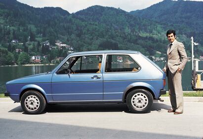 A man stands behind a blue car with a body of water and tree-covered mountains in the background.